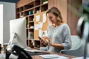 Beautiful smiling woman reading text message on mobile phone during work. Mature businesswoman checking new messages on smartphone. Casual businesswoman checking important mail while working on computer in office.