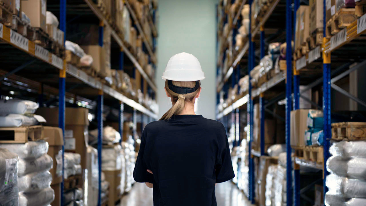 Rear view of woman standing in corridor of warehouse. She controls huge factory, wears white protective helmet and black shirt. Around her are boxes, industrial equipment and material for work in factory.